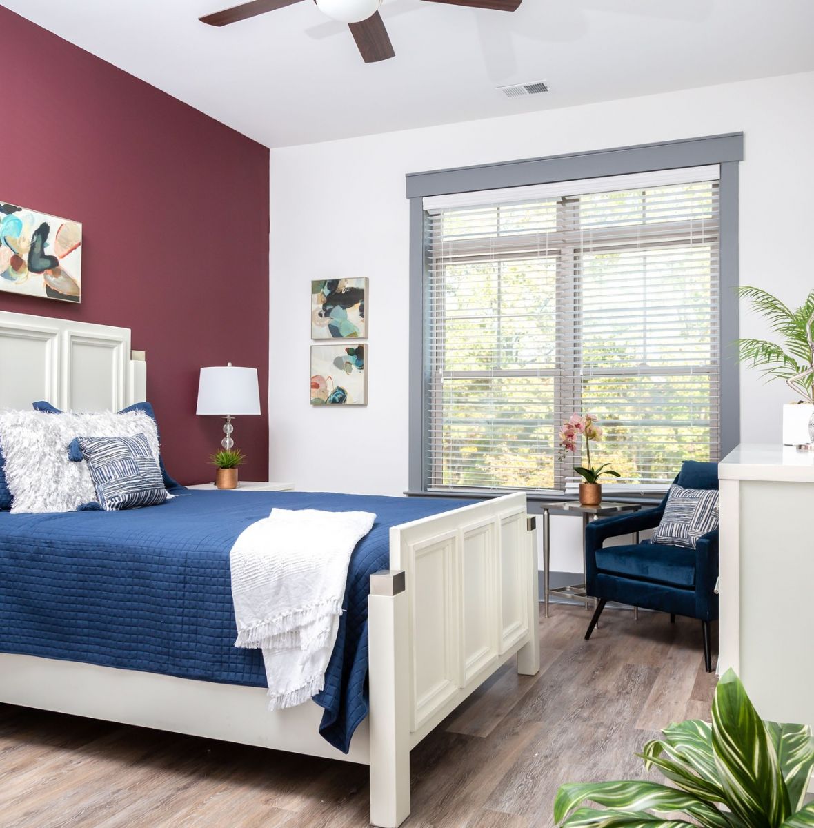 Furnished apartment bedroom with accent wall, bed, dresser, chair, and plants